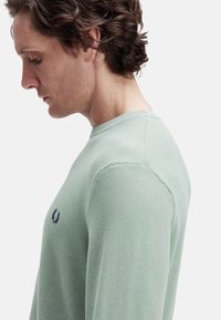Suéter de punto texturizado de color verde claro con cuello redondo y un pequeño emblema en navy en el pecho. El ajuste parece ser relajado y casual.