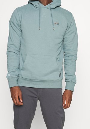 Homme portant un sweat à capuche bleu clair avec une petite broderie de vélo sur la poitrine et un pantalon gris foncé, debout sur fond blanc.
