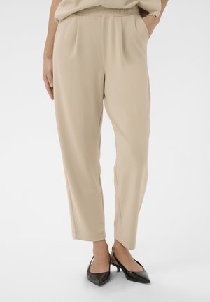 Beżowe spodnie o luźnym kroju z elastyczną talią i kieszeniami, noszone z czarnymi, ostro zakończonymi butami na obcasie typu slingback na białym tle.