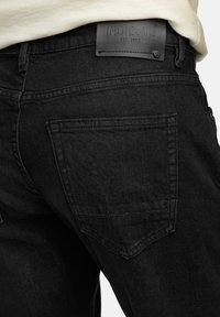 Jeans de mezclilla negra con un acabado texturizado, que muestra un pequeño bolsillo trasero y una etiqueta de marca de cuero negro que dice "INDICODE EST. 1993."