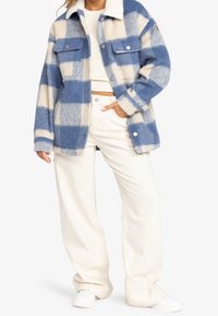 Frau in einer blau- und cremefarbigen karierten Fleecejacke, cremefarbenem Strickpullover, weißen weit geschnittenen Hosen und weißen Sneakers, steht mit einer Hand in der Tasche.