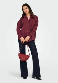 Donna che indossa una camicia button-up maroon larga e jeans scuri a zampa d'elefante, mentre tiene una borsa rossa a tracolla di fronte a uno sfondo chiaro.