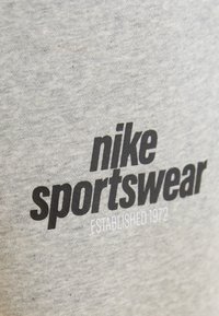 Grijze katoenen sweater met vetgedrukte zwarte tekst "nike sportswear" en "ESTABLISHED 1972" in het wit, tegen een verhoogde stofachtergrond.
