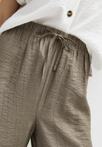 Pantalons olive verts légers et texturés avec une taille à cordon et une coupe décontractée, portés avec une chemise blanche à boutons.