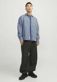 Chemise boutonnée bleu clair avec une seule poche poitrine, portée sur un t-shirt bleu foncé, associée à un pantalon noir ample et des chaussures noires.