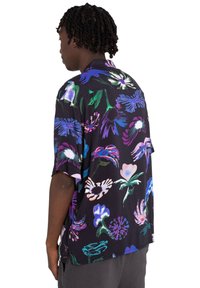 Camisa de manga corta de tejido negro ligero con patrones florales abstractos vibrantes en tonos morado, azul, rosa y verde.