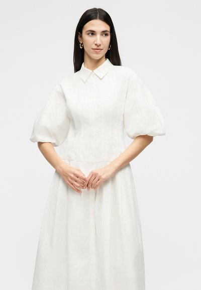 Γυναίκα με μακριά σκούρα μαλλιά, που φοράει λευκό φόρεμα με φουσκωτά μανίκια και γιακά, στέκεται με τα χέρια απαλά σταυρωμένα.