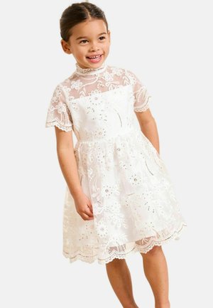 Hymyilevä nuori tyttö, joka pukeutunut valkoiseen pitsimekkoon, jossa on kukkaompeleita ja lyhyet hihat, seisoo yksinkertaista vaaleaa taustaa vasten.