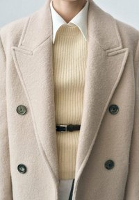 Cappotto in misto lana color nocciola con grandi bottoni, rever a punta, indossato sopra un maglione lavorato a maglia color crema, con cintura visibile. Texture liscia.
