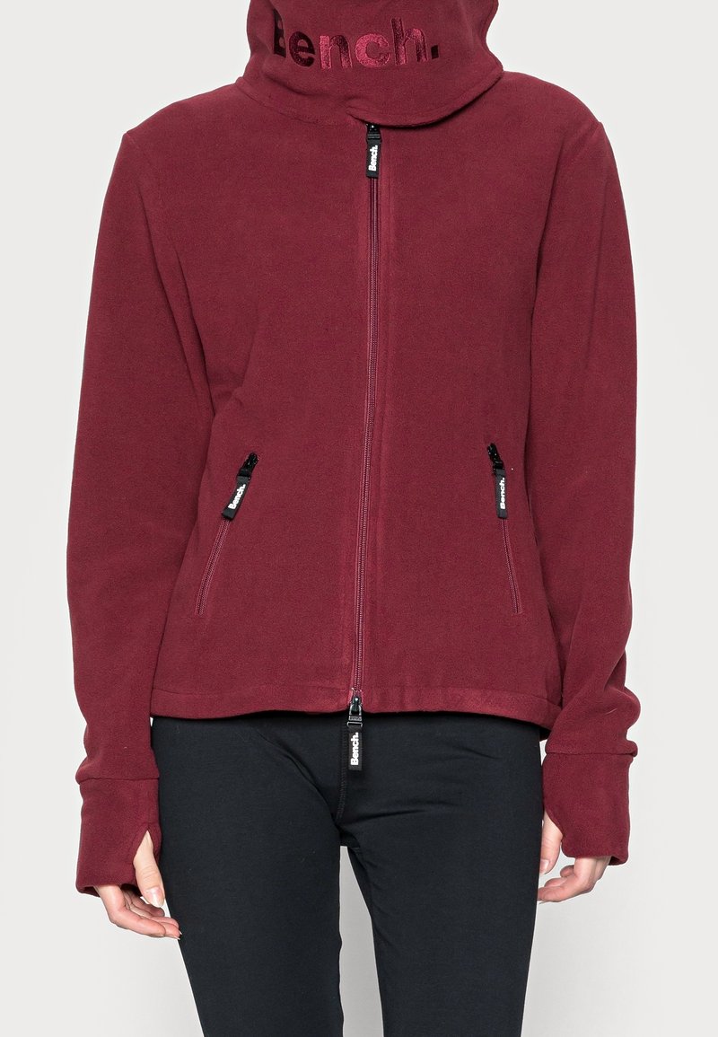 Veste polaire bordeaux zippée avec col montant, deux poches avant zippées et passages pour les pouces, portée sur un pantalon noir.