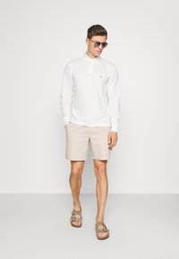 White long-sleeve polo shirt, beige linen shorts, and brown sandals. Model wearing sunglasses; simple, clean design with minimal branding.