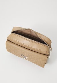 Beige Calvin Klein -laukku, jossa on rakenteellinen materiaali, vetoketjuluukku, kohotettu kuvio ja hopeinen logovihje etupuolella.