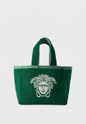 Σκούρος πράσινος τσάντα tote με ανοιχτό πράσινο περιθώριο που διαθέτει λευκό έμβλημα κεφαλής Μέδουσας στο μπροστινό μέρος και δύο κοντές λαβές.