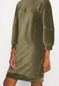 Vestido de terciopelo verde oliva con mangas largas, corte relajado, escote redondeado y textura suave, con detalles de costura sutil a lo largo de los hombros.