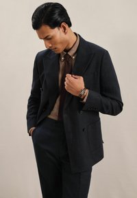 Costum bleumarin cu două rânduri de nasturi, cu o siluetă structurată, asortat cu o cămașă cu nasturi de culoare bej deschis și o cravată texturată de culoare vișinie. Ceas vizibil.