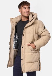 Beige dunjakke med hette, horisontale sømmer, knappestropper og glidelåslommer på sidene. Lett stoff, interiør med sølvfôr synlig.