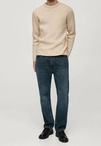 Pull beige en tricot, jean bleu à coupe droite et chaussures habillées noires. Le pull présente un col rond ; le jean a un design classique.