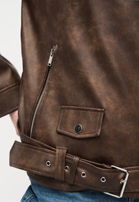 Fragment brązowej skórzanej kurtki z ukośnym srebrnym zamkiem błyskawicznym, małą kieszenią z patką i guzikiem oraz dopasowanym paskiem na tle niebieskiego denimu.