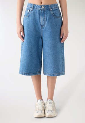 Persona che indossa pantaloncini di jeans blu di media lunghezza e scarpe da ginnastica bianche, in piedi davanti a uno sfondo chiaro e uniforme.