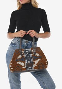 Geflochtene braune und blaue Handtasche mit geometrischem Muster, strukturiertem Design, Lederakzenten und geflochtenen Griffen, gehalten von einer Person.