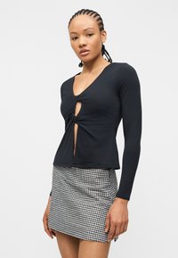 Γυναίκα με κοτσίδες, φορώντας μαύρο μακρυμάνικο τοπ με ανοίγματα μπροστά και μίνι φούστα με ασπρόμαυρο καρό σχέδιο, στέκεται μπροστά σε λευκό φόντο.