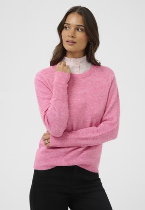 Rosa gestrickter Pullover mit langen Ärmeln und Rundhalsausschnitt, der eine weiche Textur aufweist. Getragen über einer hellen Bluse mit Rüschkragen.