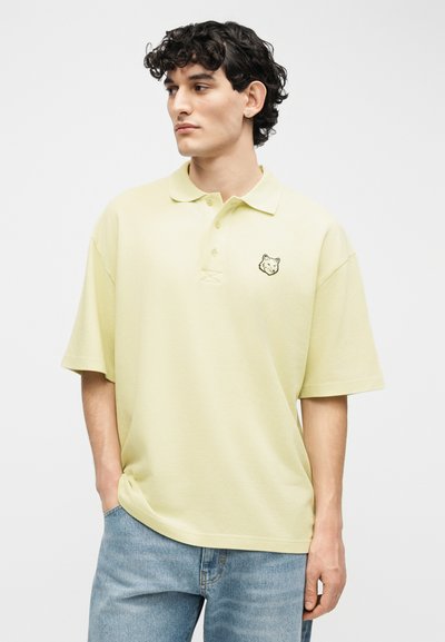 Άνδρας με σγουρά σκούρα μαλλιά που φοράει ανοιχτοκίτρινη φαρδιά μπλούζα πόλο με λογότυπο κεφαλής λύκου και ανοιχτό μπλε τζιν, κοιτάζοντας αριστερά με τα χέρια στις τσέπες.