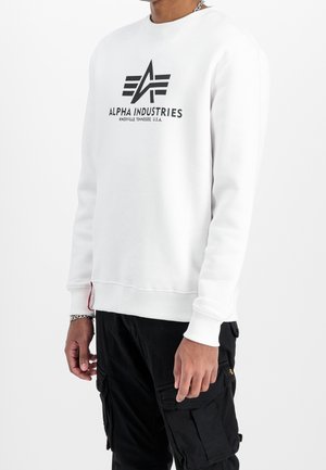 Person iført en hvid Alpha Industries sweatshirt med logo og tekst, kombineret med sorte cargobukser mod en ensfarvet hvid baggrund.