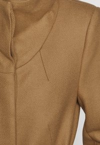 Manteau couleur tan avec une finition douce et texturée. Doté d'une capuche arrondie et de détails de couture subtils le long des épaules et du col.