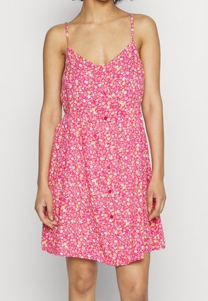 Frau trägt ärmelloses, rosa Blumenmusterkleid mit Spaghettiträgern und Knöpfen vorne, steht vor einem schlichten Hintergrund.