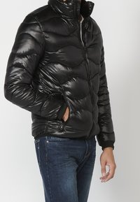 Uomo che indossa una giacca imbottita nera lucida con tasche con cerniera e jeans blu, con la mano in tasca, in piedi davanti a uno sfondo semplice.