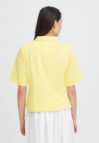 Femme aux longs cheveux noirs portant une veste jaune à manches courtes et une jupe blanche, debout de dos sur un fond clair.