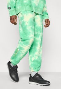 Παντελόνι φόρμας με πράσινο σχέδιο tie-dye, με απαλό υλικό και άνετη εφαρμογή, διακοσμημένο με κεντήματα στο πόδι, συνδυασμένο με μαύρα παπούτσια με κορδόνια.