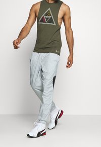 Olive green sleeveless shirt with triangular logo, paired with light gray, tapered joggers featuring black side panels and elastic cuffs. White and red sneakers.