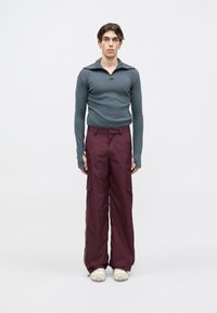 Mlada oseba stoji obrnjena naprej, oblečena v teal dolgi rokav z zadrgo in odprtinami za palce, burgundske cargo hlače in bele superge.