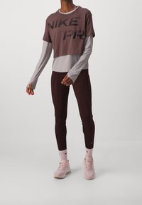 Nike lyhyttukkoinen t-paita ruskeana mustalla logolla, harmaan pitkähihasen päällä. Yhdistetty tumman burgundin värisiin leggareihin ja vaaleanpunaisiin urheilujalkineisiin.