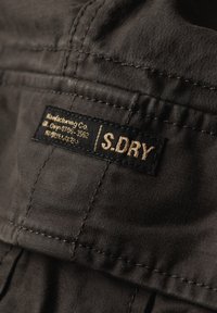 Tissu noir avec une étiquette noire cousue affichant le nom de la marque S.DRY et un texte additionnel en jaune.