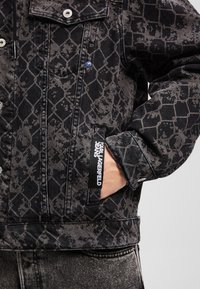 Giacca di jeans nera con un motivo testurizzato, hardware argentato e un'etichetta con logo. Il design include una tasca sul petto e dettagli distintivi.