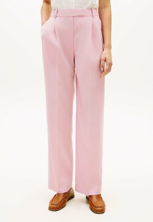 Person iført lyserøde bukser med høj talje og vide ben samt brune loafers, stående med hænderne afslappet ned langs siden på en ensfarvet baggrund.