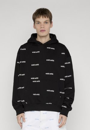 Mladý muž oblečený v čiernej mikine s kapucňou a bielych nohaviciach, oblečenie je pokryté opakujúcimi sa textovými logami „eckō unltd.“ na jednoduchom pozadí.