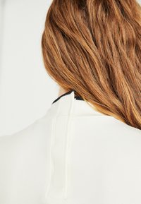 Red hair falling over the back of a white high-collar garment with a vertical seam and a small fabric tab at the neckline.