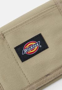 Khaki stofflommebok med en rektangulær svart etikett med hvit og rød "Dickies"-logo, sydd med synlige sømmer langs kantene.