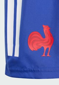 Červený stylizovaný emblém kohouta na modré látce se dvěma vertikálními bílými pruhy, pravděpodobně součást sportovního oděvu nebo dresu.