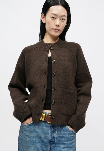 Cardigã de lã castanha com gola redonda, seis botões na frente e dois bolsos laterais. Combinado com uma camisola preta e jeans de ganga.