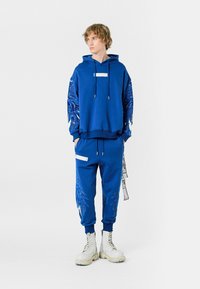 Sudadera azul con diseños gráficos de alas en las mangas, combinada con pantalones jogger azul con acentos laterales y herrajes blancos. Usada con botas blancas.