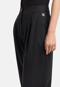 Pantaloni neri con una texture liscia, caratterizzati da una vita alta, pieghe e un piccolo accento in metallo vicino alla tasca posteriore.