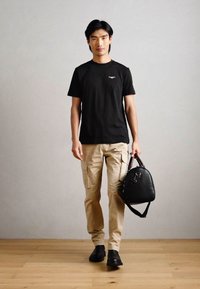 Melna kokvilnas T-krekls, beigtu krāsu bikses ar sānu kabatām, melna ādas soma ar rokturi un melni ikdienas apavi uz koka grīdas pelēkas sienas fonā.