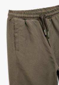 Khaki dresowe spodnie z elastycznym pasem, regulowanym sznurkiem i bocznymi kieszeniami. Charakteryzują się gładką fakturą i subtelnym detalem w stębnowaniu.