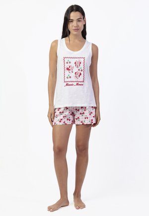 Mujer con top blanco sin mangas con caras de Minnie Mouse y diseños de cerezas, combinada con pantalones cortos rosas con patrón de cerezas, de pie descalza.