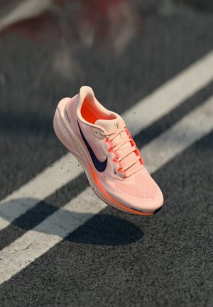 Chaussure de course Nike rose clair avec un swoosh noir flottant au-dessus d'une route asphaltée avec des lignes blanches.